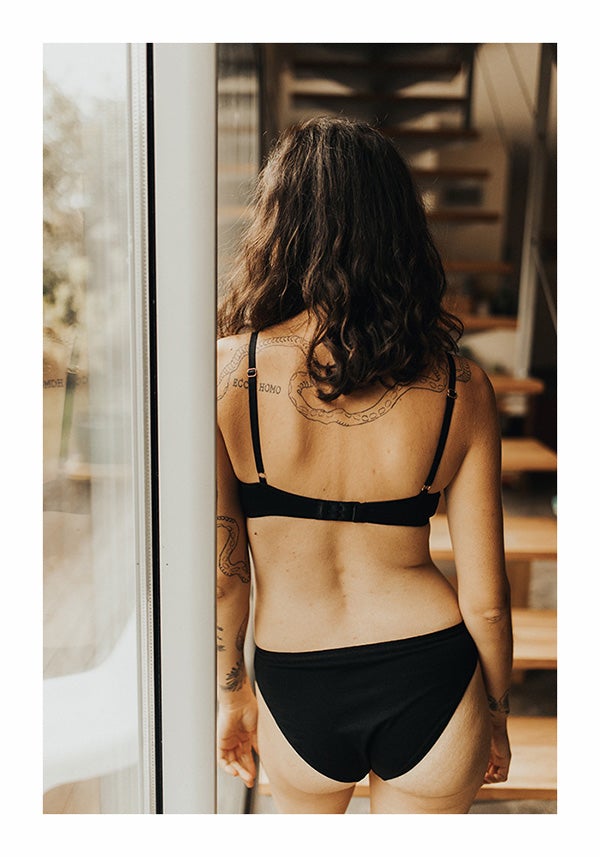Femme debout de dos, devant des escaliers, portant une culotte et un soutien-gorge noirs