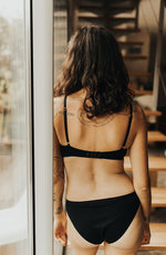 Femme de dos devant des escaliers, habillée avec une culotte et un soutien-gorge noirs