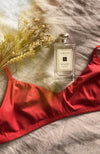 Soutien-gorge sans armature en coton bio rouge terracotta, posé sur un lit avec une bouteille de parfum et un bouquet de fleurs séchées