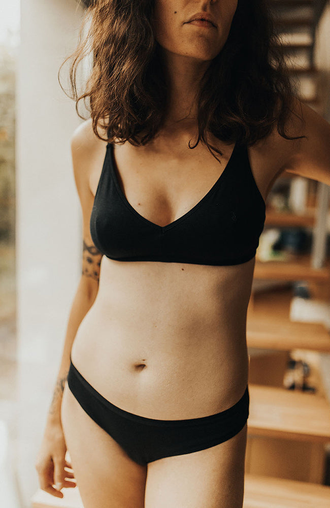 Femme debout devant des escaliers, portant un soutien-gorge sans armature, forme triangle, et une culotte taille basse en coton bio noir